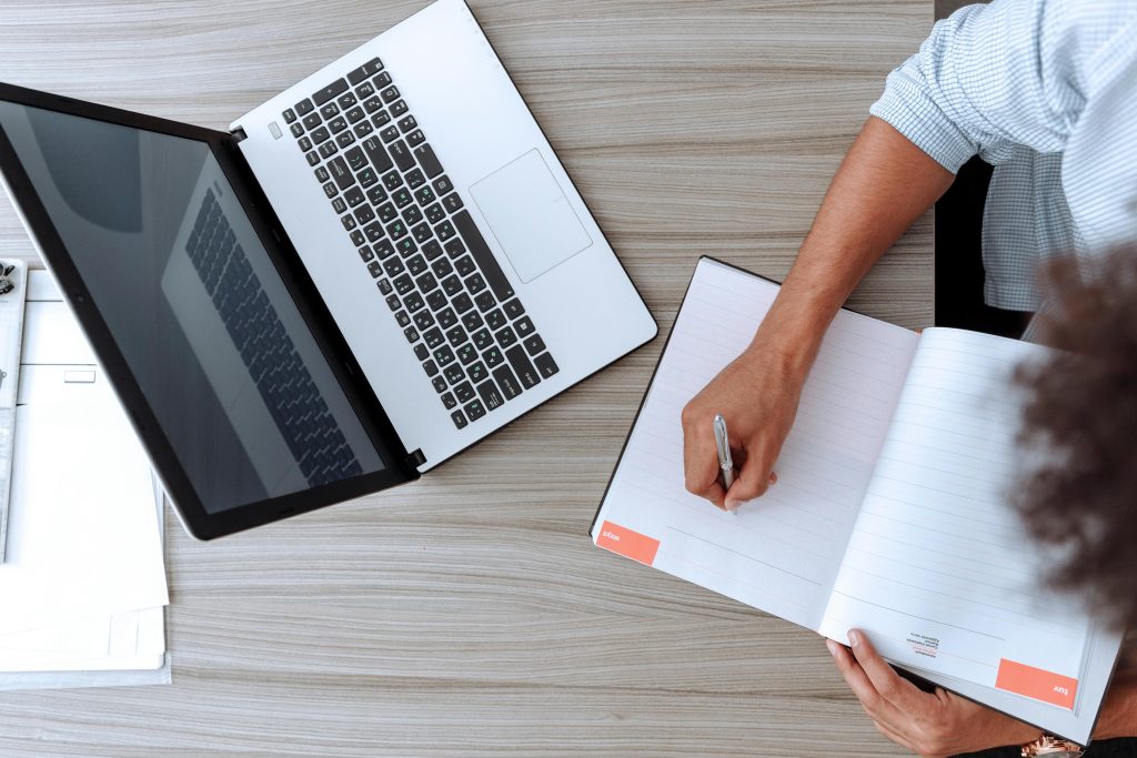 Person writing notes at desk with a laptop and open notebook, capturing a modern workspace.